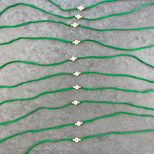 May include: A collection of ten thin green cord bracelets, each featuring a single white circular bead with the letter V printed in gold. Each bead is flanked by two small gold-toned metallic spacers, arranged in parallel rows across the frame.