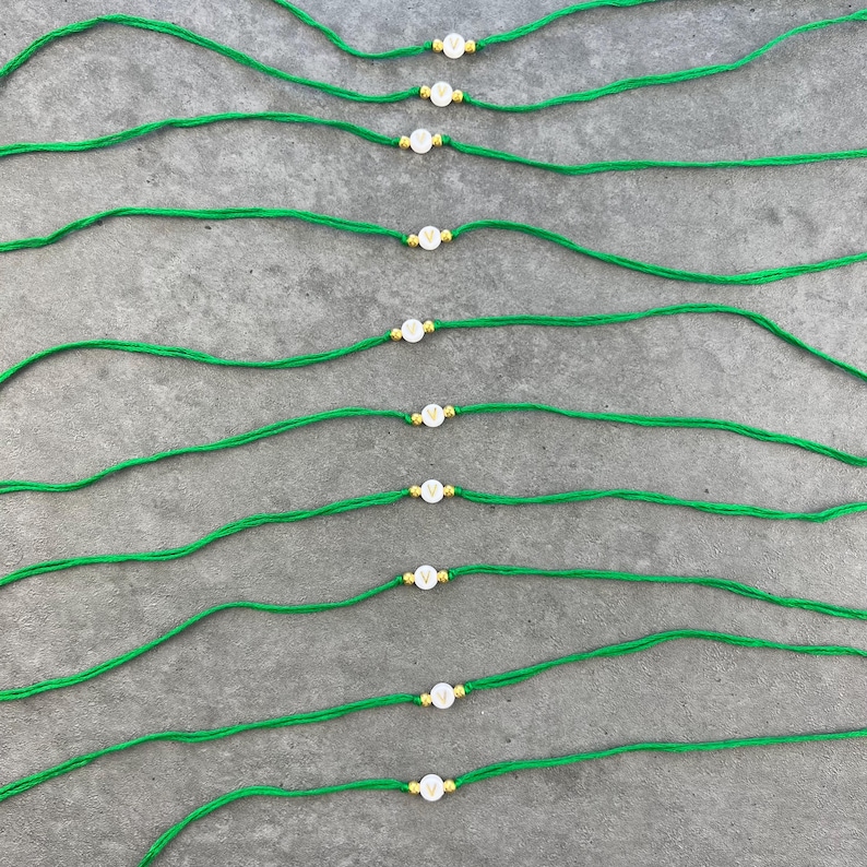 May include: A collection of ten thin green cord bracelets, each featuring a single white circular bead with the letter V printed in gold. Each bead is flanked by two small gold-toned metallic spacers, arranged in parallel rows across the frame.