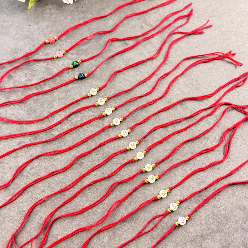 May include: A collection of thin red string bracelets, each featuring a single round bead with a letter printed on it. Some beads are white with black letters, while others are colored with white letters, all accented by small gold-toned metal beads.