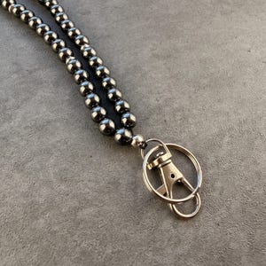 K&ouml;nnte beinhalten: Ein Schl&uuml;sselband mit zwei Str&auml;ngen aus glatten, reflektierenden, dunkelgrauen Metallperlen. Die Perlen sind aufgereiht und mit einem silberfarbenen Metall-Karabinerhaken sowie einem runden Schl&uuml;sselring zum Befestigen von Schl&uuml;sseln oder Ausweisen verbunden.