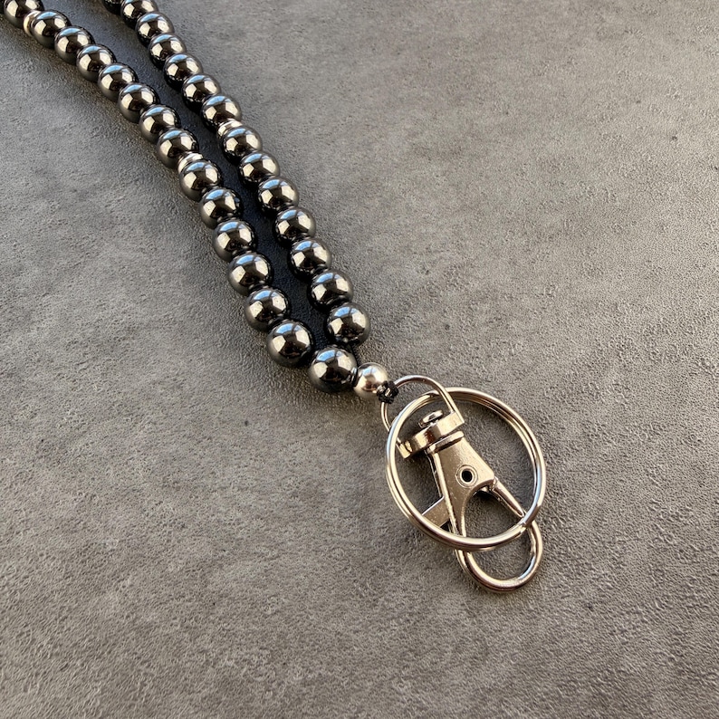 K&ouml;nnte beinhalten: Ein Schl&uuml;sselband mit zwei Str&auml;ngen aus glatten, reflektierenden, dunkelgrauen Metallperlen. Die Perlen sind aufgereiht und mit einem silberfarbenen Metall-Karabinerhaken sowie einem runden Schl&uuml;sselring zum Befestigen von Schl&uuml;sseln oder Ausweisen verbunden.