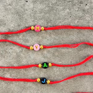 May include: Four red string bracelets, each featuring a single round letter bead flanked by two small gold-tone metallic beads. The top two bracelets have pink beads with yellow letters N and Y, while the bottom two have black beads with green A and blue J.