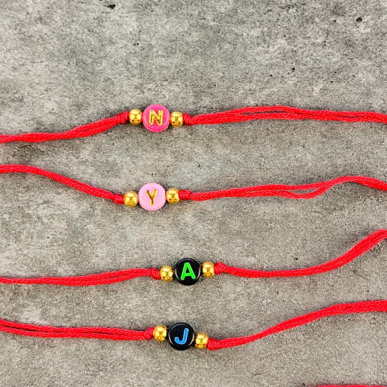 May include: Four red string bracelets, each featuring a single round letter bead flanked by two small gold-tone metallic beads. The top two bracelets have pink beads with yellow letters N and Y, while the bottom two have black beads with green A and blue J.