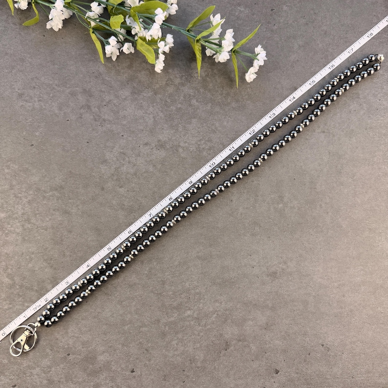 K&ouml;nnte beinhalten: Ein langes Schl&uuml;sselband aus dunkelgrauen, metallisch gl&auml;nzenden, kugelf&ouml;rmigen Perlen mit einem silberfarbenen Karabinerverschluss an einem Ende. Ein wei&szlig;es Ma&szlig;band liegt neben dem Band und zeigt eine L&auml;nge von etwa 46 cm an.