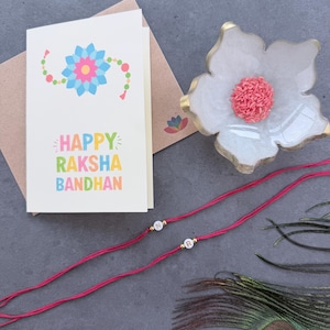 May include: A white greeting card with a colorful floral illustration and the text Happy Raksha Bandhan, a white flower-shaped dish containing pink rice grains, and two red thread bracelets featuring white letter beads N and R.