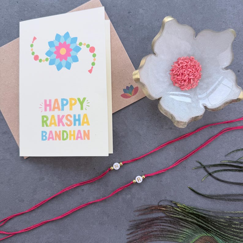 May include: A white greeting card with a colorful floral illustration and the text Happy Raksha Bandhan, a white flower-shaped dish containing pink rice grains, and two red thread bracelets featuring white letter beads N and R.