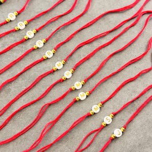 May include: A collection of red string bracelets, each featuring a single white circular bead with a gold letter and two small gold-toned spacer beads on either side. The letters visible on the beads include R, M, S, and J, arranged in a row.