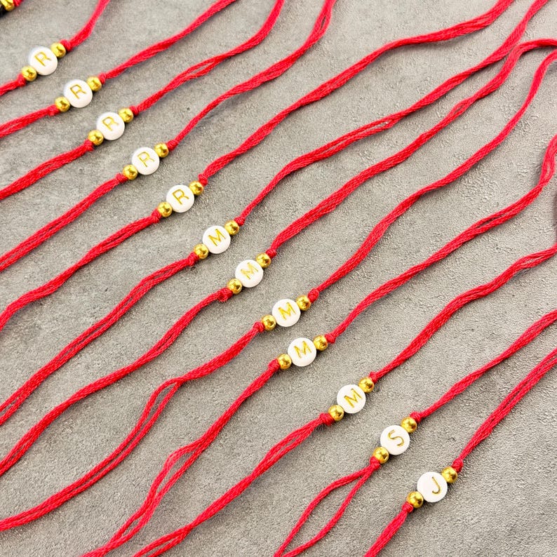 May include: A collection of red string bracelets, each featuring a single white circular bead with a gold letter and two small gold-toned spacer beads on either side. The letters visible on the beads include R, M, S, and J, arranged in a row.