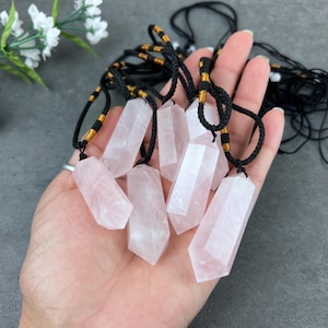 May include: A group of seven rose quartz crystal point pendants on black cords with gold beads. The pendants are all different sizes and shapes, but they are all polished and have a smooth, shiny finish.