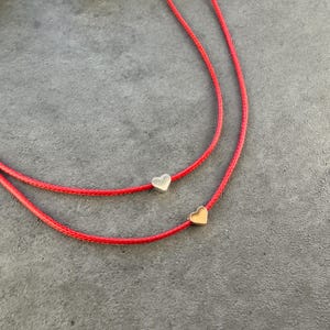 May include: Two red cord necklaces, each with a small heart pendant. One heart is silver, the other is gold. The necklaces are displayed on a gray surface. The jewelry is simple and elegant.