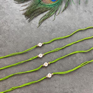 May include: Four lime green string bracelets, each featuring a single white circular bead with a gold letter. Two bracelets display the letter V and two display the letter A, with small gold-toned metallic spacer beads flanking each letter bead.