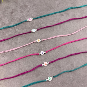 May include: Six thin cord bracelets in teal, dark purple, light pink, bright pink, magenta, and teal. Each bracelet features a single white circular bead with a colored letter and two small metallic silver spacer beads on either side of the letter.