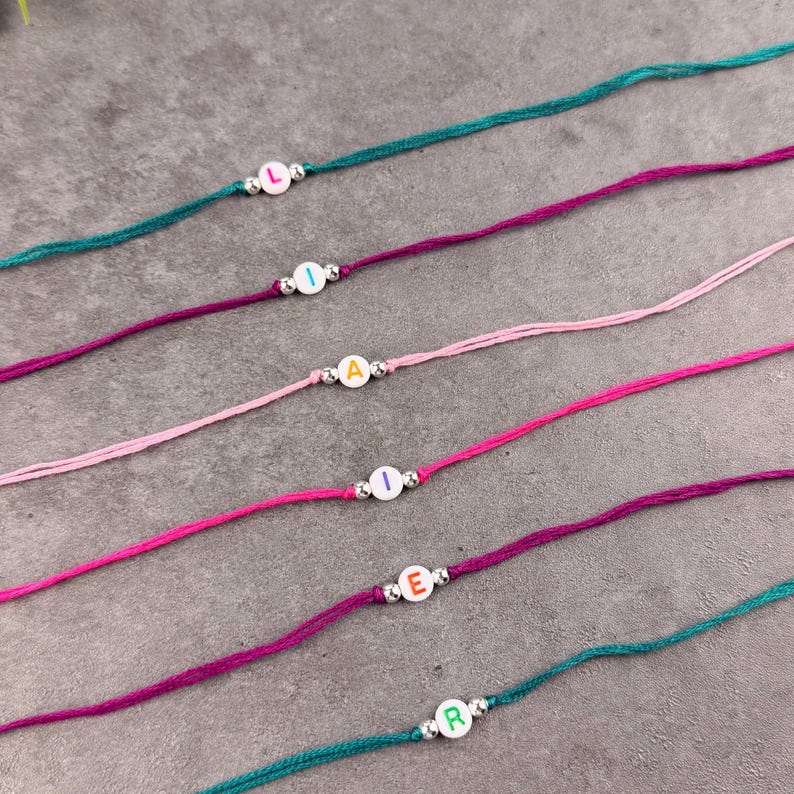May include: Six thin cord bracelets in teal, dark purple, light pink, bright pink, magenta, and teal. Each bracelet features a single white circular bead with a colored letter and two small metallic silver spacer beads on either side of the letter.