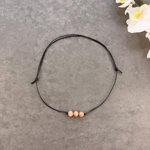 May include: A black cord necklace featuring three peach-colored beads. The necklace is simple and elegant, with adjustable knots on each end. The beads are evenly spaced along the cord, creating a minimalist design.