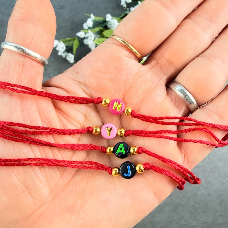 May include: Four red string bracelets, each featuring a single round bead with a letter in the center. The beads are pink with a gold N, light pink with a gold Y, black with a green A, and black with a blue J. Small gold-toned beads flank each letter bead.