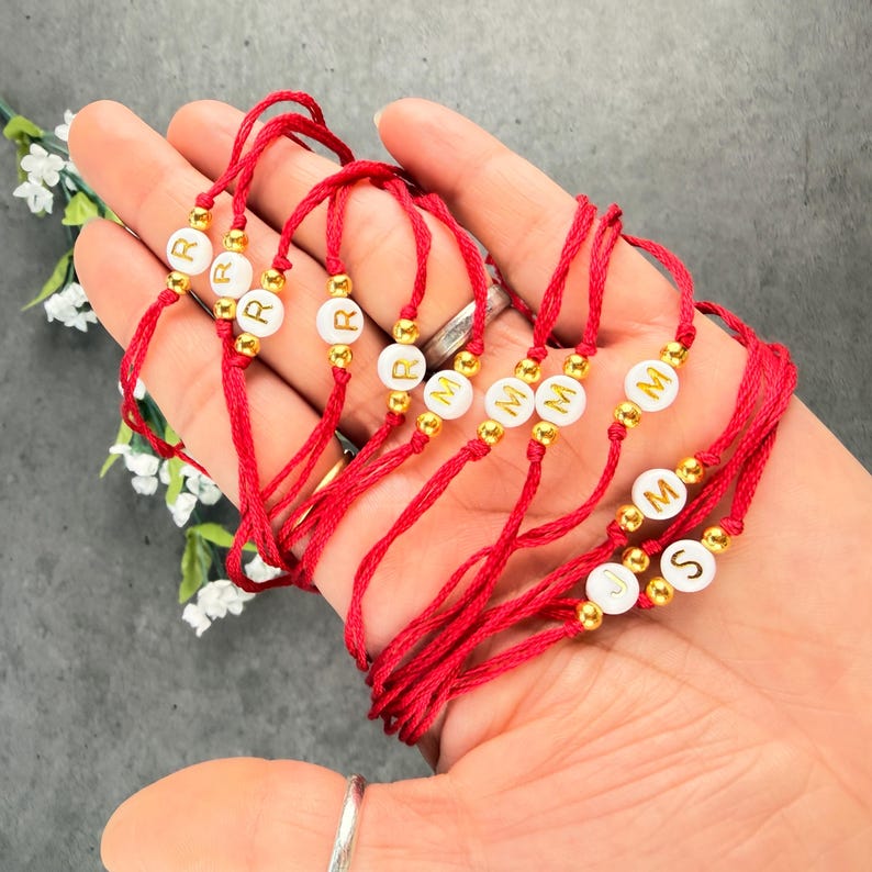 May include: A collection of red string bracelets featuring white circular beads with gold-colored letters and small gold-colored spacer beads. The bracelets display various letters including R, M, J, and S, arranged in a simple, adjustable design.