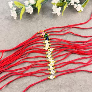May include: A collection of red string bracelets, each featuring a single round bead with a letter printed on it, separated by small gold-toned metallic spacer beads. The letters include N, Y, A, J, R, M, S, and J, arranged in a vertical line.