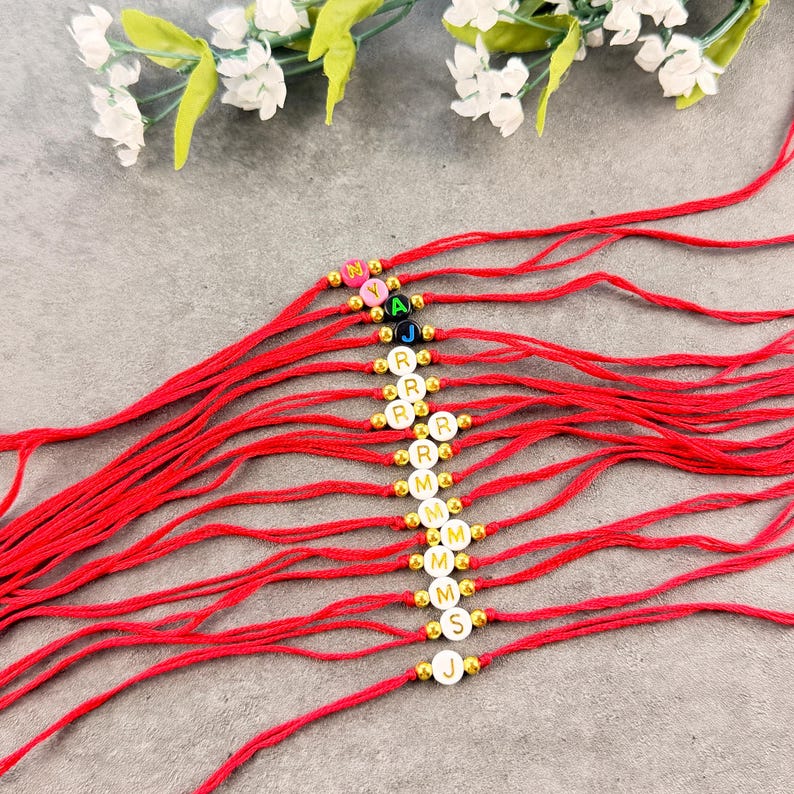 May include: A collection of red string bracelets, each featuring a single round bead with a letter printed on it, separated by small gold-toned metallic spacer beads. The letters include N, Y, A, J, R, M, S, and J, arranged in a vertical line.