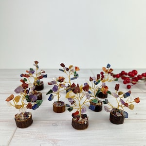 May include: A collection of small decorative gemstone trees. Each tree features a gold wire trunk and branches adorned with colorful, irregularly shaped gemstones. The trees are set in small, round, bark-covered bases, creating a natural aesthetic.