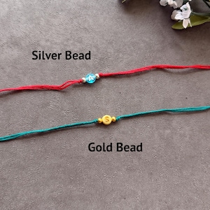 May include: Two string bracelets. The top bracelet is red with a blue bead featuring the white letter A, flanked by two silver beads. The bottom bracelet is teal with a yellow bead featuring the white letter S, flanked by two gold beads.