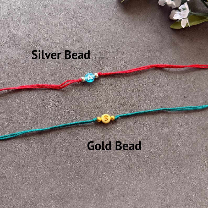 May include: Two string bracelets. The top bracelet is red with a blue bead featuring the white letter A, flanked by two silver beads. The bottom bracelet is teal with a yellow bead featuring the white letter S, flanked by two gold beads.