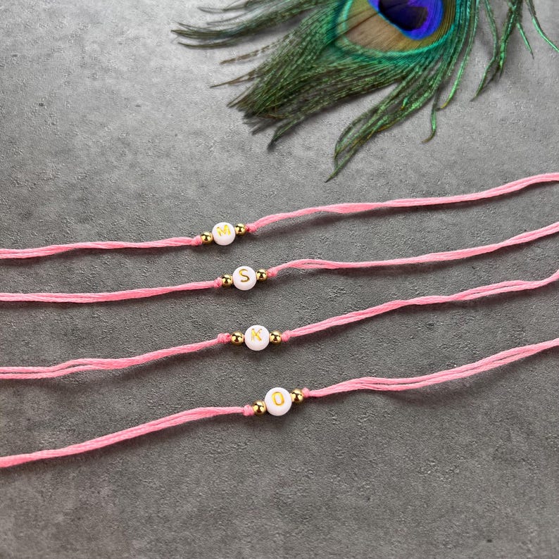 May include: Four pink cord bracelets, each featuring a single white round bead with a gold letter. The letters displayed are M, S, K, and O. Each bead is flanked by two small gold-toned metallic spacer beads on the thin pink string material.