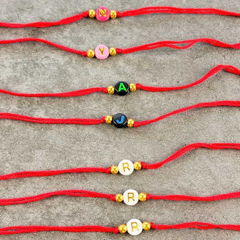 May include: Seven red string bracelets, each featuring a single letter bead flanked by two small gold-toned metallic beads. The letter beads include pink with N, pink with Y, black with A, black with J, white with R, white with R, and white with R.