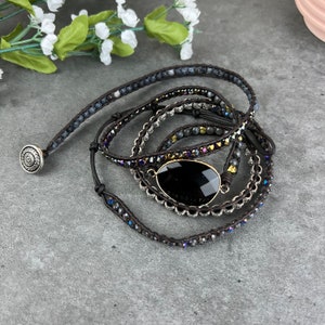 May include: A multi-strand wrap bracelet with a large black gemstone pendant. The bracelet is made of brown leather cord, silver beads, and dark gray stones.