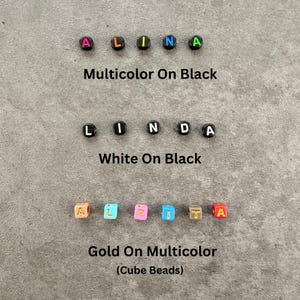 May include: Three rows of alphabet beads. The first row spells ALINA with multicolored letters on black beads. The second row spells LINDA with white letters on black beads. The third row spells ALPITA with gold letters on multicolored cube beads.