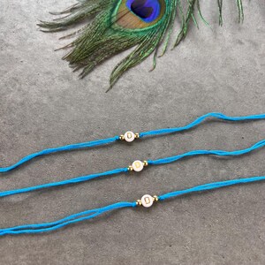 May include: Three bright blue cord bracelets, each featuring a single white circular bead with the letter D printed in gold, flanked by two small gold-toned metallic spheres. The bracelets are arranged horizontally across the frame.