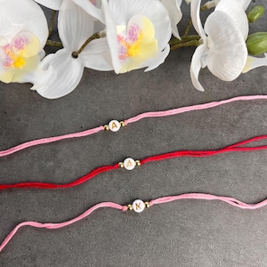 May include: Three personalized bracelets with initial beads. The bracelets are made of pink and red string, each featuring a gold-colored bead with a letter. White orchids are in the background. The letters are A, A, and K.