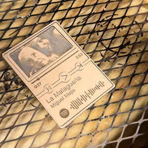 May include: A wooden music player with a black and white photo of a person. The player has a title that reads "La Malagueña" by "Miguel Mejía" and a Spotify logo. The player has a timer that reads "0:17" and "3:53".