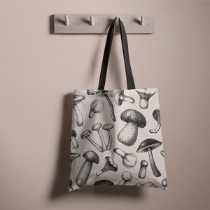 May include: A white tote bag with black handles and a black and white mushroom print. The bag is hanging on a hook.