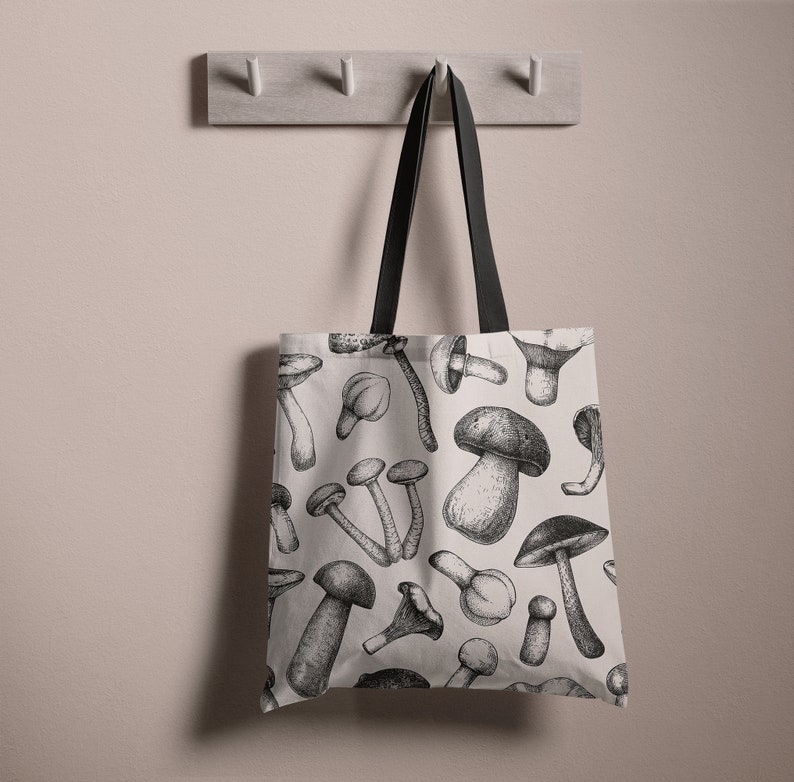 May include: A white tote bag with black handles and a black and white mushroom print. The bag is hanging on a hook.