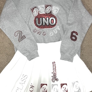 May include: A gray sweatshirt and white pleated skirt set. The sweatshirt features a red and silver "UNO" design with "2026" and "UNO OUT" text. The skirt has "CLASS OF Yohana" and "2026" card graphics.