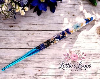 May include: A blue and white crochet hook with embedded flowers and gold flakes. The hook is made of clear resin and has a blue handle. The hook is on a wooden surface.