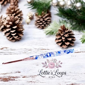 May include: A rose gold crochet hook with a blue and white glitter handle. The handle has a snowflake design. The hook is on a white wooden surface with pine cones and greenery.