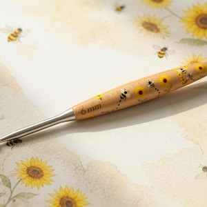 May include: A 6 mm crochet hook with a light wood handle featuring a printed pattern of yellow sunflowers and small bees. The hook has a smooth silver metal tip and a tapered shaft, with the size 6 mm clearly engraved on the wooden handle.