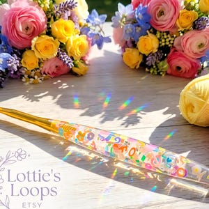 May include: A transparent crochet hook with a gold-coloured hook end and a handle filled with colourful shapes and letters. The hook is on a light surface, with flower bouquets and yarn balls in the background. The text "Lottie's Loops" is visible.