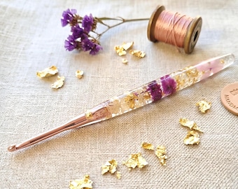 May include: A rose gold crochet hook with a clear resin handle containing embedded purple dried flowers and gold leaf flakes. The hook is marked with 5.5mm. Nearby are a wooden spool of rose gold thread and scattered gold leaf flakes.