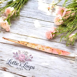 May include: A rose gold crochet hook with pink and white pressed flowers encased in clear resin. The hook is on a white wooden surface with pink flowers in the background. The text "Lottie's Loops Etsy" is visible in the lower left corner of the image.