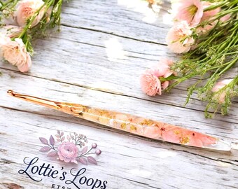 May include: A rose gold crochet hook with pink and white pressed flowers encased in clear resin. The hook is on a white wooden surface with pink flowers in the background. The text "Lottie's Loops Etsy" is visible in the lower left corner of the image.