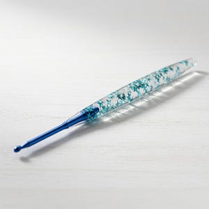 May include: A crochet hook featuring a solid blue metal tip and a clear, tapered handle filled with small, teal-coloured star-shaped glitter. The handle is transparent, allowing the internal star decorations to be clearly visible throughout the body.