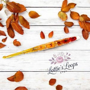 May include: A gold crochet hook with a clear handle filled with pressed flowers and leaves. The handle is decorated with orange, yellow, and red flowers. The hook is lying on a white wooden surface with autumn leaves scattered around it. The text "Lottie's Loops Etsy" is visible in the lower right corner of the image.