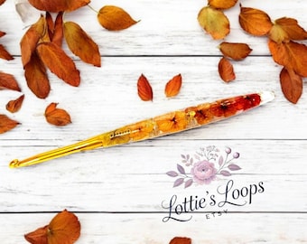 May include: A gold crochet hook with a clear handle filled with pressed flowers and leaves. The handle is decorated with orange, yellow, and red flowers. The hook is lying on a white wooden surface with fall leaves scattered around it. The text "Lottie's Loops Etsy" is visible in the lower right corner of the image.