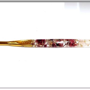 Könnte beinhalten: Eine goldene Häkelnadel mit einem durchsichtigen Griff, der mit gepressten rosa und weißen Blumen gefüllt ist.