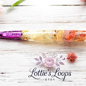 May include: A purple and gold crochet hook with a clear handle filled with dried flowers. The handle is made of resin and has a floral design. The hook is 20 cm long.