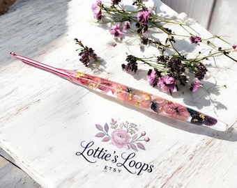May include: A pink crochet hook with dried flowers and gold flakes embedded in the clear resin handle. The hook is lying on a white wooden surface with a floral design and the text "Lottie's Loops Etsy".