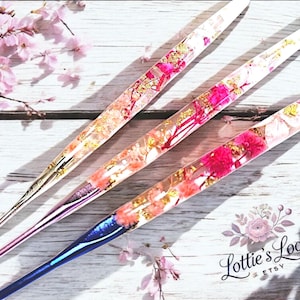 May include: Three colourful crochet hooks with embedded flowers and gold flakes. The hooks are clear, pink and blue. The hooks are laid out on a white wooden surface.