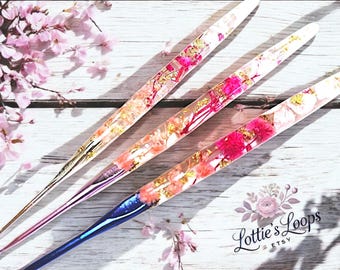 May include: Three colorful crochet hooks with embedded flowers and gold flakes. The hooks are clear, pink, and blue. The hooks are laid out on a white wooden surface.
